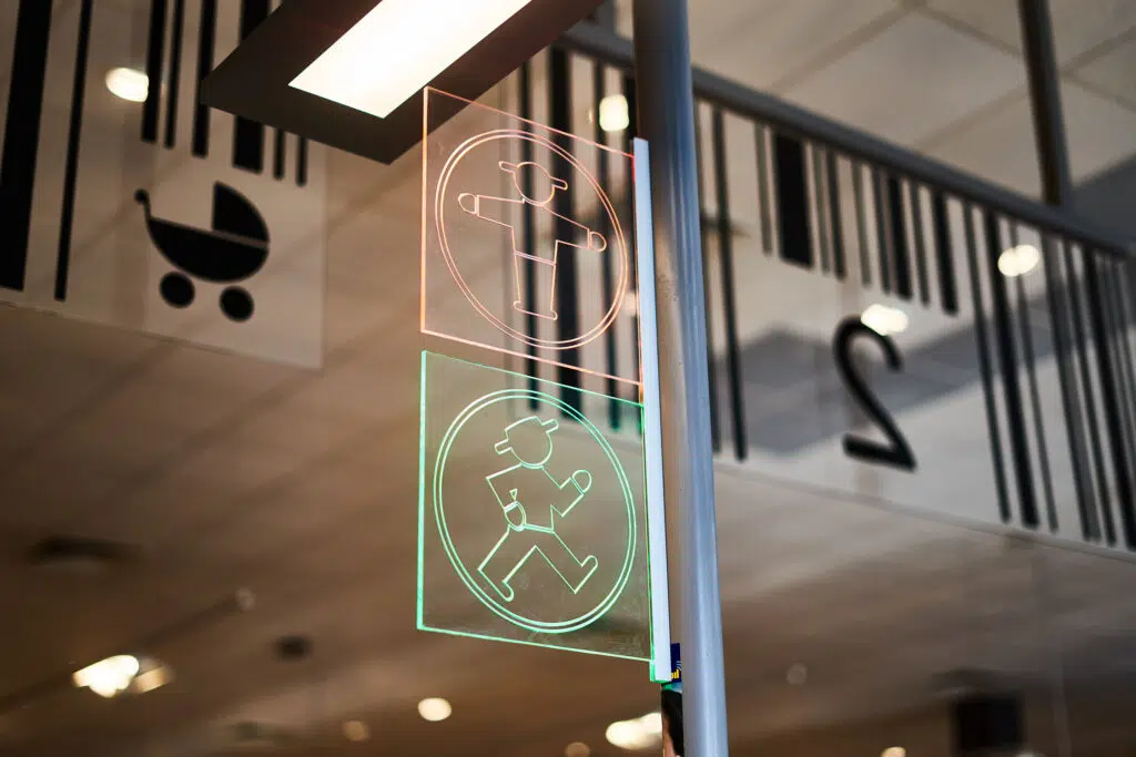 Zwei beleuchtete Glasschilder mit Ampelmännchen-Symbolen in Rot (stehend) und Grün (gehend). Im Hintergrund eine Glaswand mit Barcodes, der Zahl „2“ und einem Kinderwagensymbol. Die moderne Umgebung ist gut beleuchtet und spiegelt ein stilvolles Design wider.