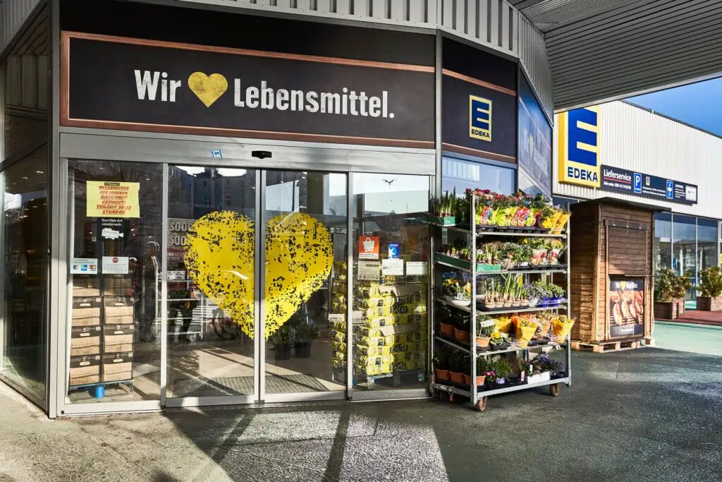 Das Bild zeigt den Eingang eines EDEKA-Supermarkts. Über den automatischen Glastüren befindet sich ein großes Schild mit der Aufschrift „Wir [HERZ] Lebensmittel.“ Ein großes gelbes Herz ist auf die Glasscheibe gedruckt. Rechts vom Eingang steht ein Pflanzenregal mit Blumen und Kräutern.
