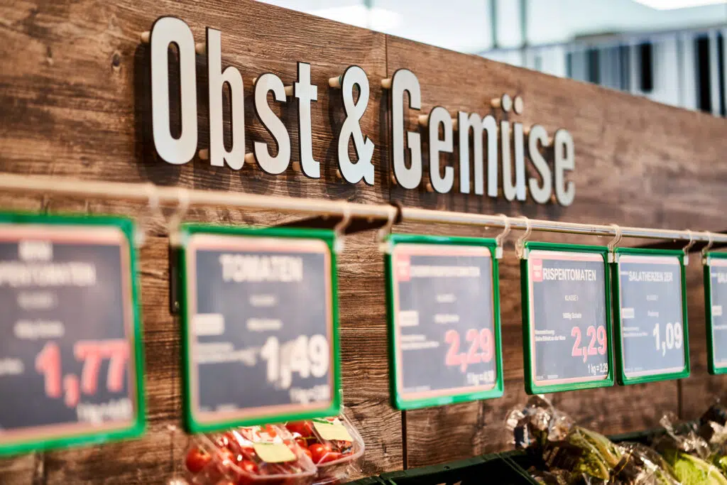 Das Bild zeigt eine Obst- und Gemüseabteilung in einem Supermarkt. Im Fokus steht ein Holzschild mit dem Schriftzug „Obst & Gemüse“ in großen, plastischen Buchstaben. Darunter sind Preisschilder mit grünen Rahmen an einer Schiene befestigt, die verschiedene Produkte auszeichnen.