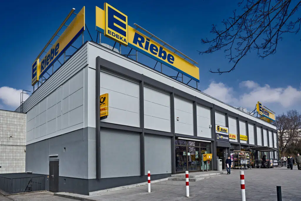 Das Bild zeigt die Außenansicht eines EDEKA-Marktes mit dem Namen „Riebe“. Auf dem Dach befinden sich große gelb-blaue Schilder mit dem EDEKA-Logo und dem Schriftzug „Riebe“. Die Fassade ist grau mit abgesetzten dunklen Rahmen um die Fenster. Neben dem Eingang sind eine Postfiliale, ein Lotto-Geschäft und ein Bereich mit Einkaufskörben und Pflanzen zu sehen. Einige Personen befinden sich vor dem Geschäft.