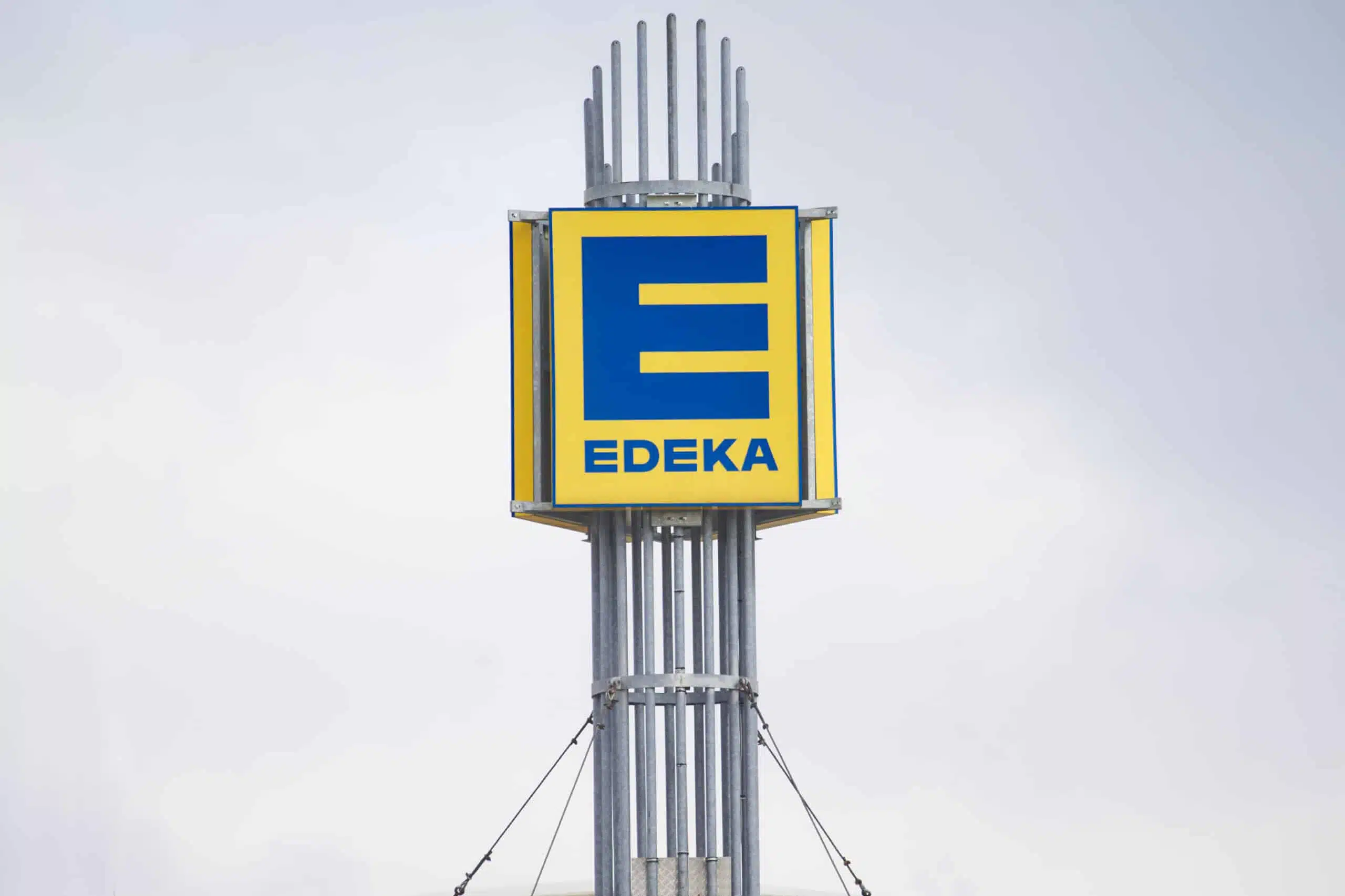 Das Bild zeigt ein großes EDEKA-Logo auf einem hohen, stabilen Metallmast. Das quadratische Schild ist gelb mit einem blauen "E" und dem Schriftzug "EDEKA" darunter. Der Mast besteht aus mehreren senkrechten Metallrohren und ist mit Stahlseilen abgesichert. Der Hintergrund ist ein grauer, bewölkter Himmel.