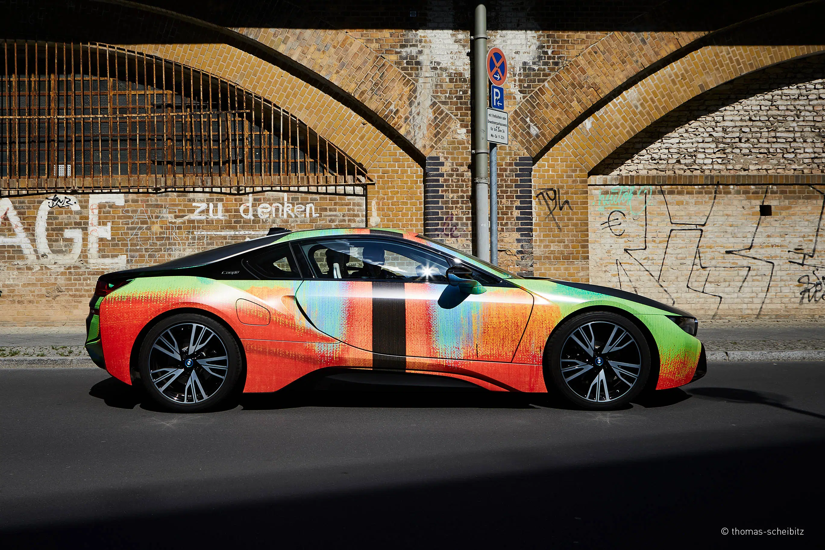 Dieses Bild zeigt einen seitlich aufgenommenen BMW i8 mit einer auffälligen, kunstvollen Fahrzeugfolierung. Das Design besteht aus leuchtenden Farben und abstrakten Mustern, die dem Wagen eine einzigartige Optik verleihen. Im Hintergrund ist eine Backsteinwand mit Graffiti und ein Durchfahrtsverbotsschild zu sehen, was einen urbanen Kontrast zur modernen und kreativen Gestaltung des Fahrzeugs bildet.