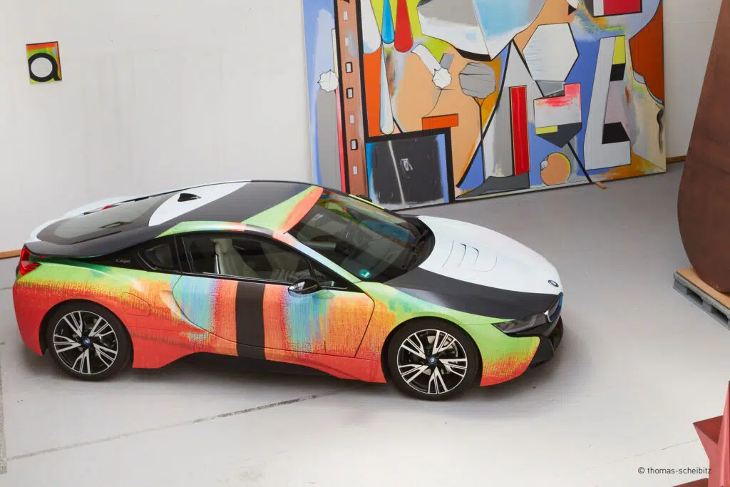 Dieses Bild zeigt einen BMW i8 mit einer einzigartigen, kunstvollen Fahrzeugfolierung, die an moderne Malerei erinnert. Der Wagen steht in einem Atelier oder einer Galerie, im Hintergrund sind abstrakte Kunstwerke zu sehen, die das kreative Design ergänzen.