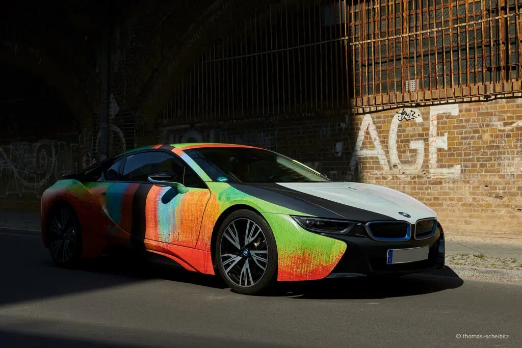 Dieses Bild zeigt einen BMW i8 mit einer auffälligen, kunstvollen Fahrzeugfolierung, die farbenfrohe abstrakte Muster aufweist. Das Auto steht in einer urbanen Umgebung vor einer Backsteinwand mit Graffiti, was den Kontrast zwischen moderner Kunst und Street-Art betont.