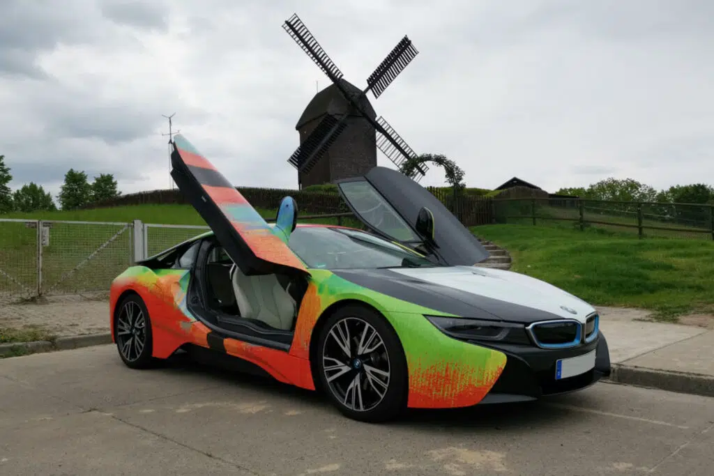 Dieses Bild zeigt einen folierten BMW i8 mit auffälligem, farbenfrohem Design und geöffneten Flügeltüren. Im Hintergrund steht eine traditionelle Windmühle, die dem modernen Fahrzeug einen interessanten Kontrast verleiht.
