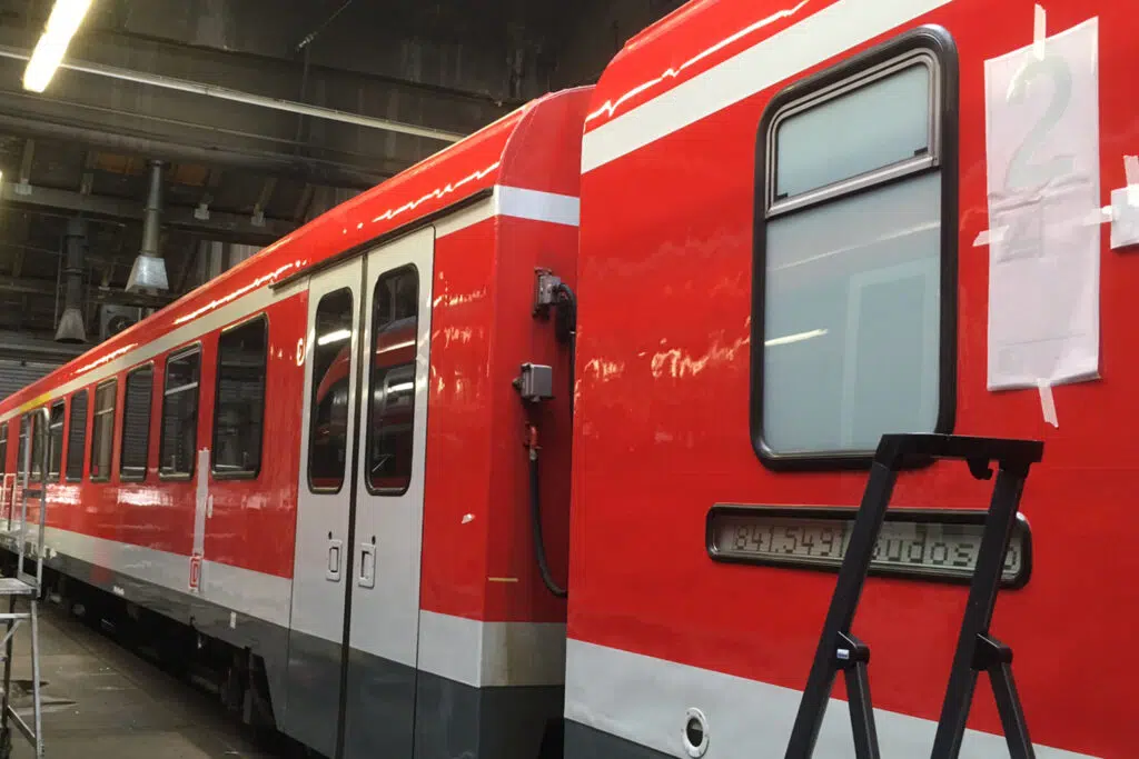 Das Bild zeigt einen roten Schienenwagen mit einer frischen Folierung in einer Werkstatthalle. Die weiße Linienführung ist bereits angebracht, und eine Leiter steht an der Seite des Fahrzeugs. An der Wand ist eine Vorlage für eine Kennzeichnung befestigt.