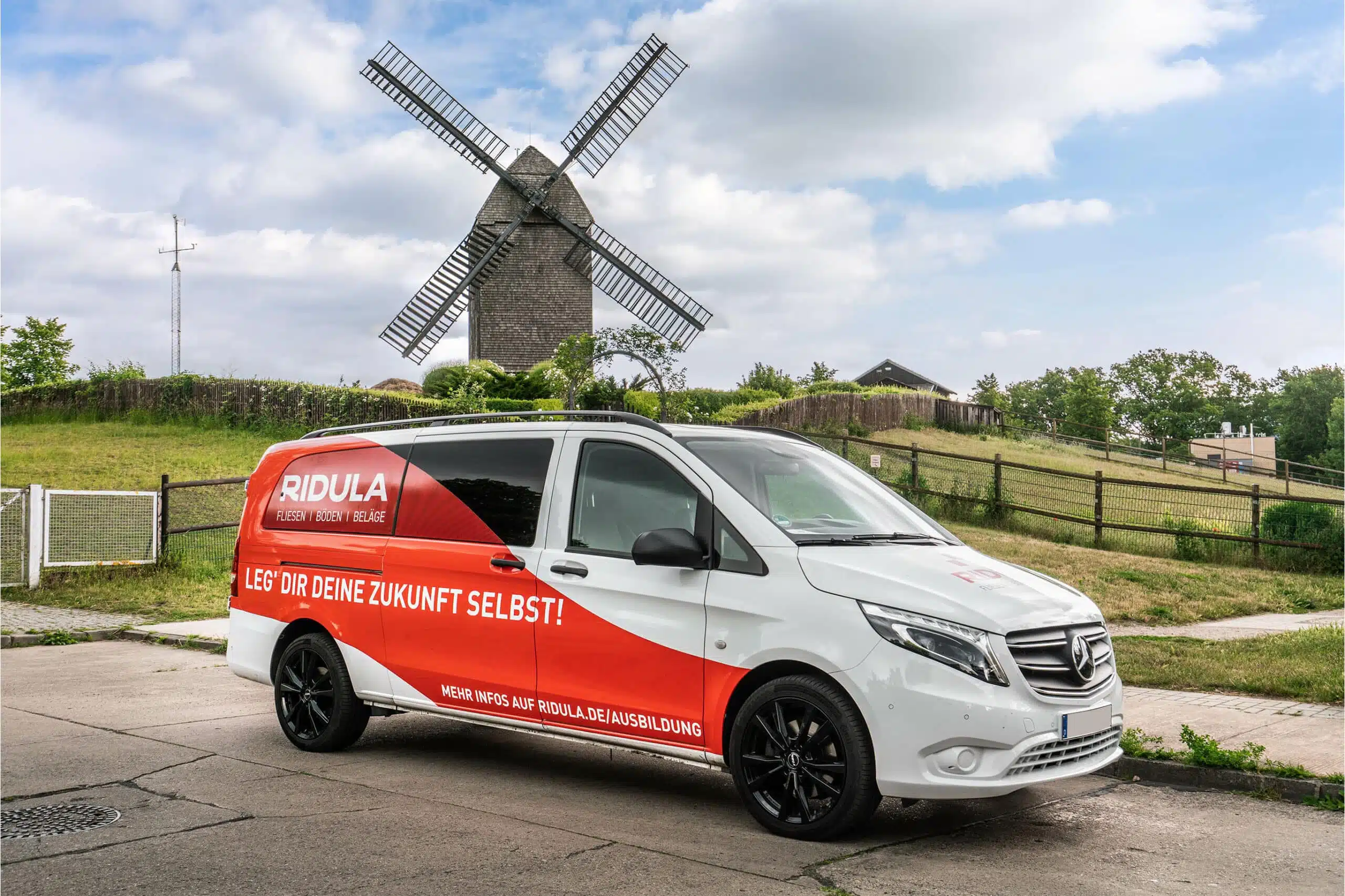 Ein rot-weiß folierter Mercedes-Transporter von "RIDULA" mit Werbebeschriftung steht vor einer historischen Windmühle. Der Slogan "Leg’ dir deine Zukunft selbst!" sowie die Webadresse für weitere Informationen sind sichtbar.