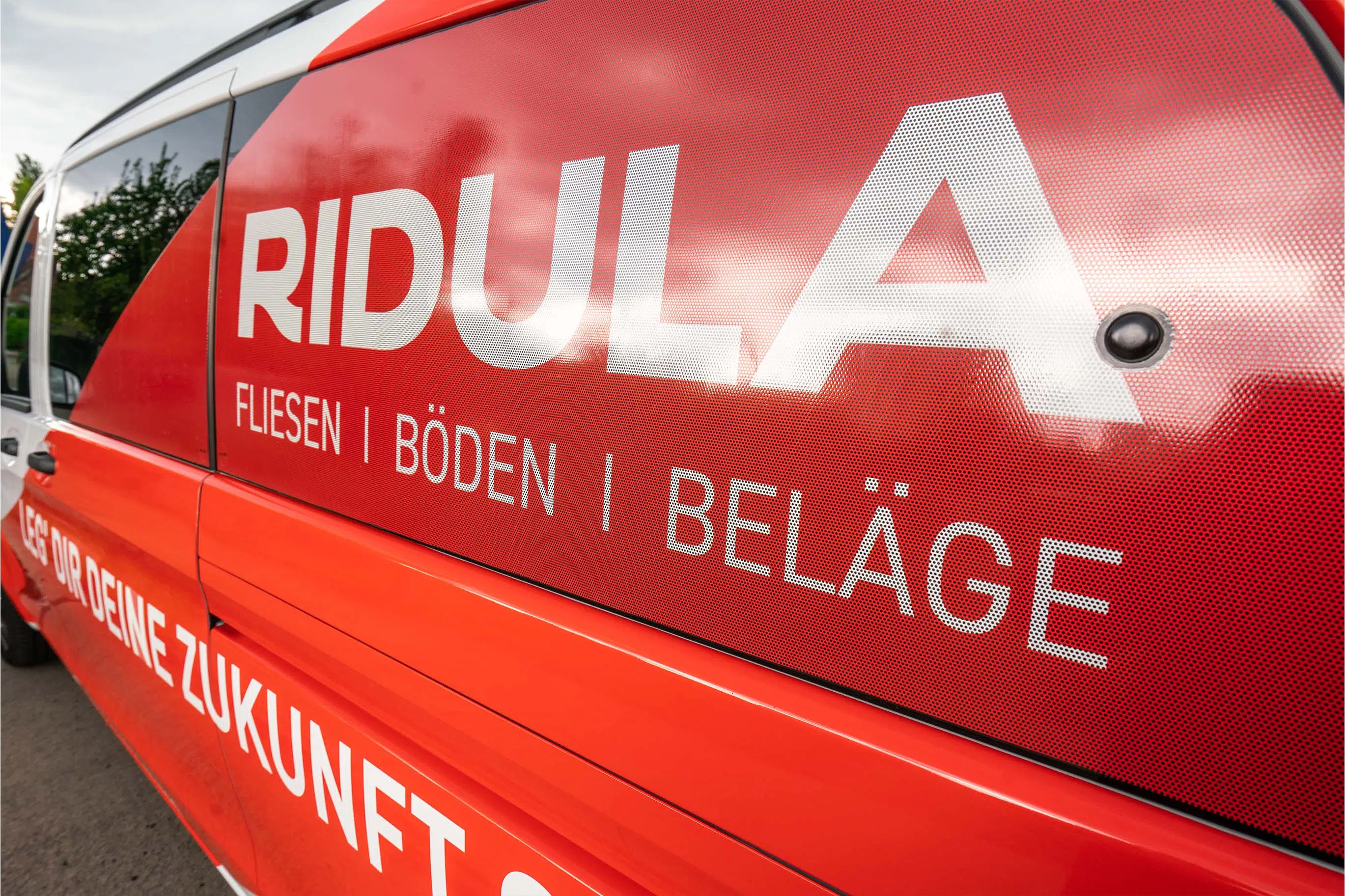 Nahaufnahme eines rot-weißen Firmenfahrzeugs mit Werbebeschriftung für "RIDULA" – spezialisiert auf Fliesen, Böden und Beläge. Der Slogan "Leg dir deine Zukunft" ist ebenfalls sichtbar. Das Fenster ist mit Lochfolie beklebt.