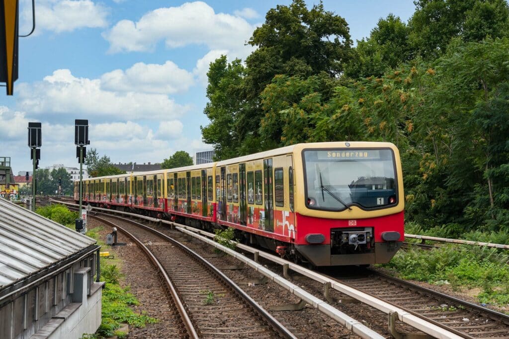 Sonderzug der Berliner Ringbahn mit Jubiläums-Design auf freier Strecke. Der Zug trägt eine spezielle Folierung mit historischen Motiven und ist in den klassischen Farben der S-Bahn Berlin gehalten. Die Umgebung ist grün mit klarem Himmel.