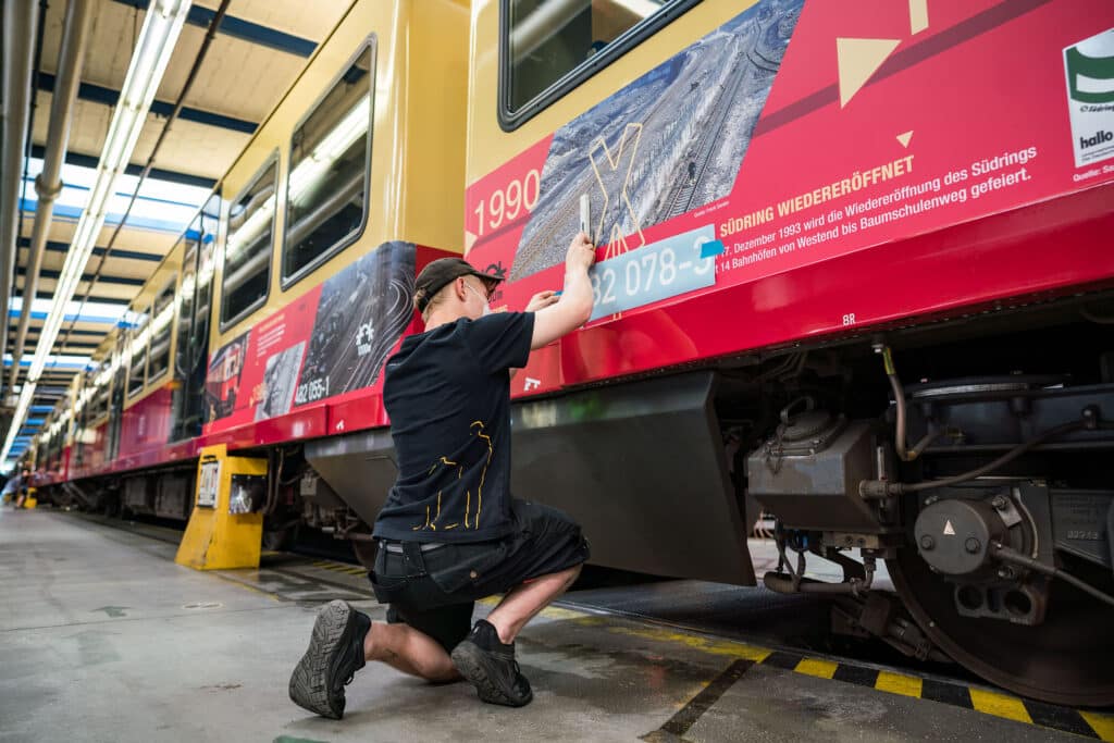 Ein Mitarbeiter bringt präzise eine historische Beklebung an einer Berliner Ringbahn an. Die Folie zeigt Informationen zur Wiedereröffnung des Südrings und historische Bilder, passend zum Jubiläum der Ringbahn.