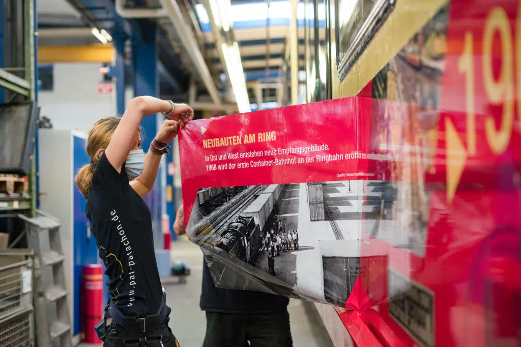Eine Mitarbeiterin bringt eine großflächige historische Folierung auf einer Berliner Ringbahn an. Die Beklebung zeigt Informationen zur Geschichte der Ringbahn und wird präzise aufgetragen.