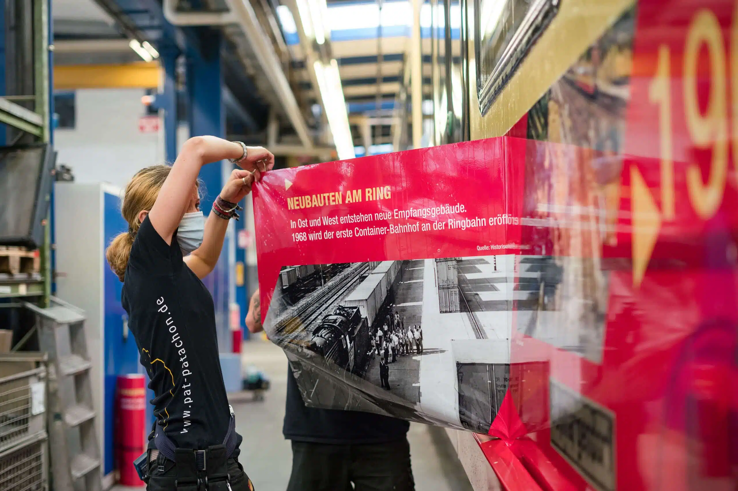 Eine Mitarbeiterin bringt eine großflächige historische Folierung auf einer Berliner Ringbahn an. Die Beklebung zeigt Informationen zur Geschichte der Ringbahn und wird präzise aufgetragen.