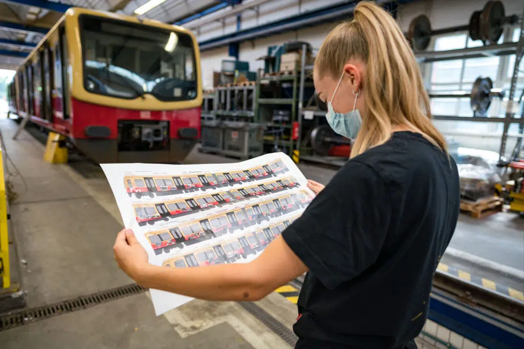 Eine Mitarbeiterin prüft den Designentwurf für die Jubiläumsfolierung der Berliner Ringbahn. Im Hintergrund steht eine S-Bahn bereit für die Beklebung in einer Werkstatt mit technischer Ausstattung.