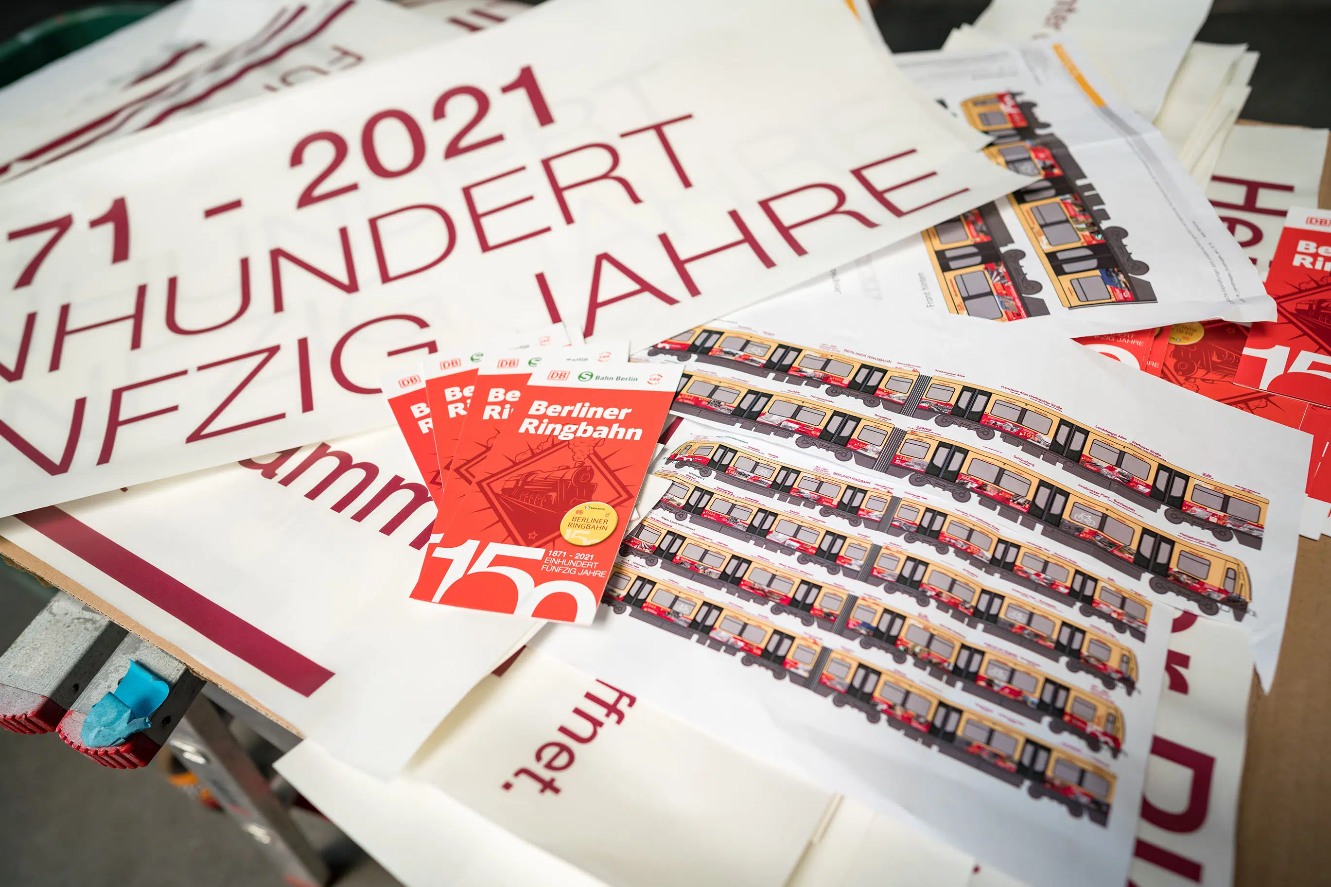 Druckvorlagen und Layouts für das Jubiläumsdesign der Berliner Ringbahn. Auf dem Tisch liegen Folien, Skizzen und Fahrplanhefte mit dem S-Bahn-Design zum 150-jährigen Bestehen, inklusive historischer Elemente und moderner Gestaltung.