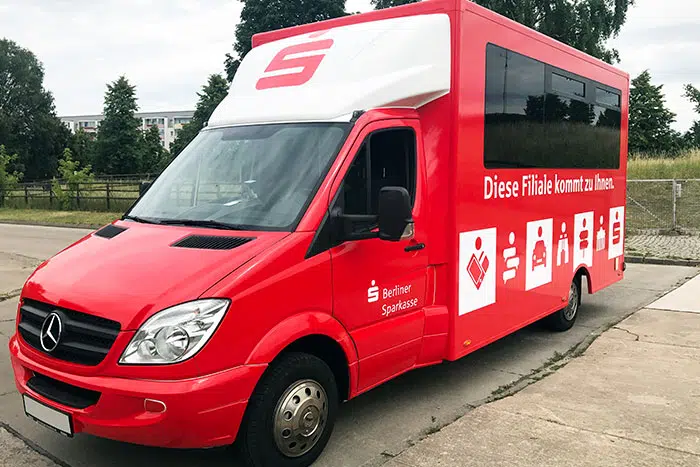 Ein roter Mercedes-Benz Transporter der Berliner Sparkasse, umgebaut als mobile Filiale. Auf der Seite steht „Diese Filiale kommt zu Ihnen.“ mit Sparkassen-Logos und Symbolen für Bankdienstleistungen. Der Bus steht auf einer Straße mit Grünflächen im Hintergrund.