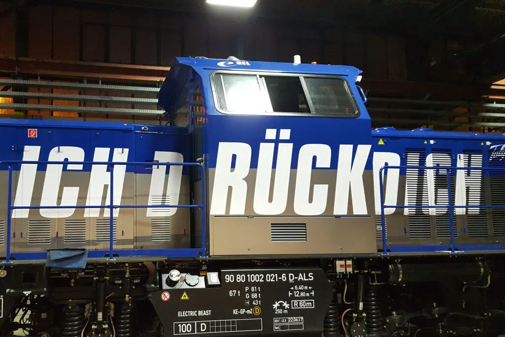 Das Bild zeigt eine blau-beige Lokomotive mit großflächigem weißen Schriftzug. Der Text ist auf die Seitenflächen verteilt und teilweise verdeckt. Technische Beschriftungen und Sicherheitskennzeichnungen befinden sich an der Unterseite der Lok.