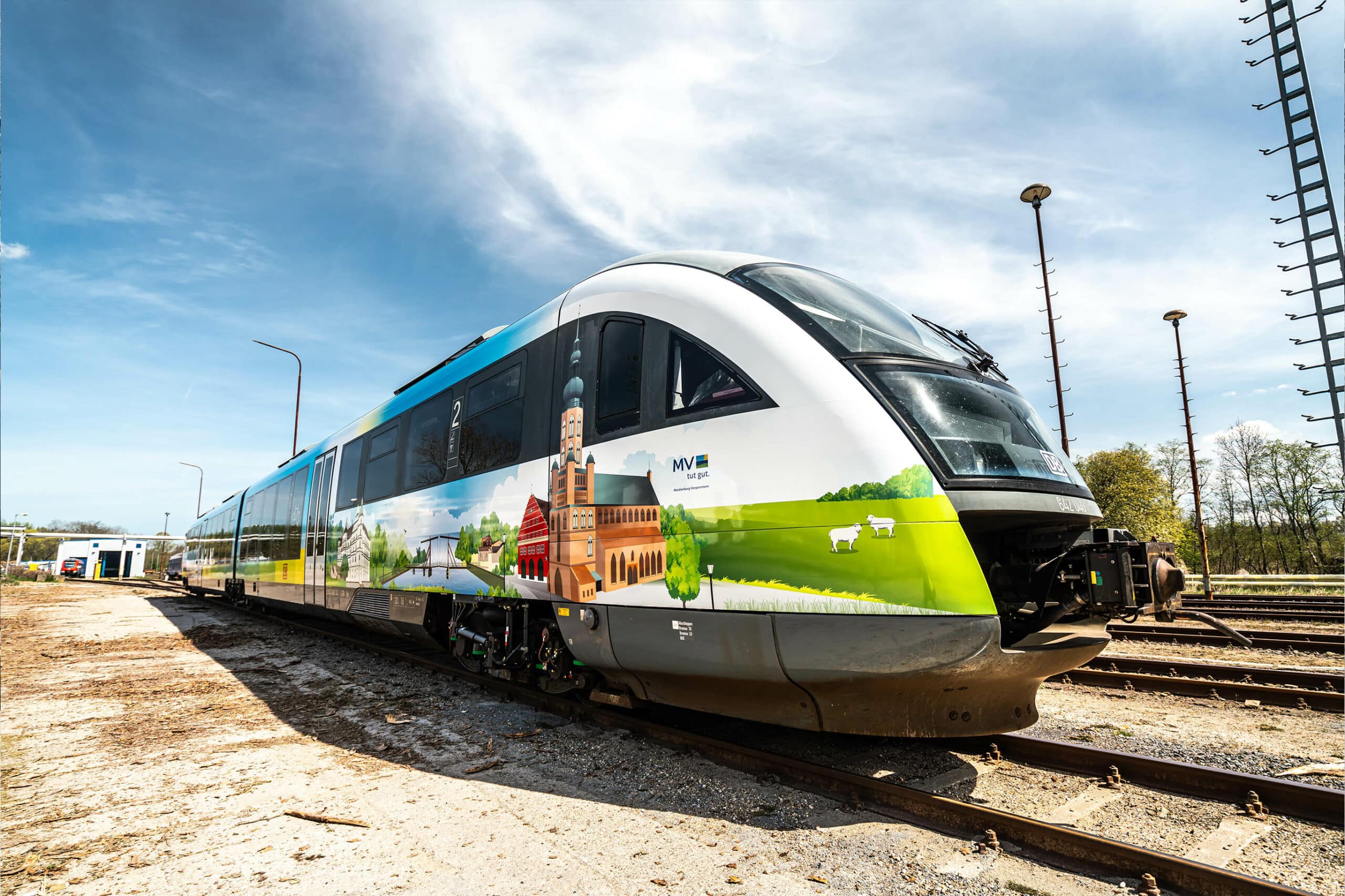 Ein modern folierter Zug mit regionalen Usedom-Motiven steht auf den Gleisen. Die Gestaltung zeigt Sehenswürdigkeiten, grüne Landschaften und kulturelle Elemente der Region, während der Himmel klar und blau über dem Zug erstrahlt.
