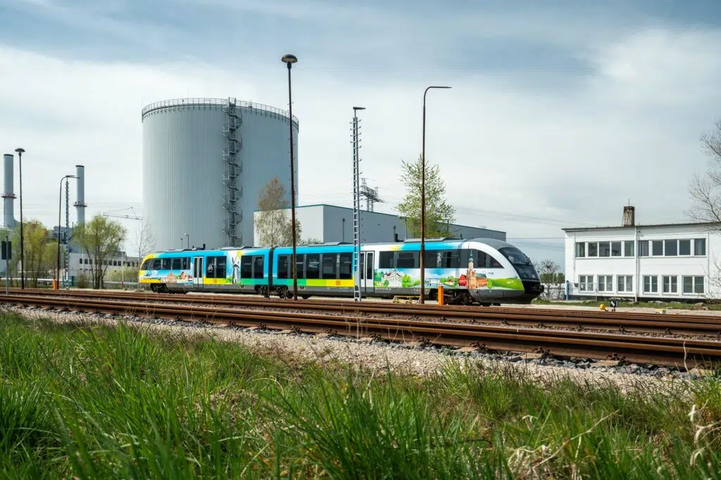 Ein modern gestalteter Zug mit Usedom-Motiven fährt durch eine industrielle Umgebung. Die bunte Folierung zeigt regionale Sehenswürdigkeiten, darunter eine Windmühle und eine Altstadt, und verbindet Natur und Stadtbild auf einer harmonischen Gestaltung.