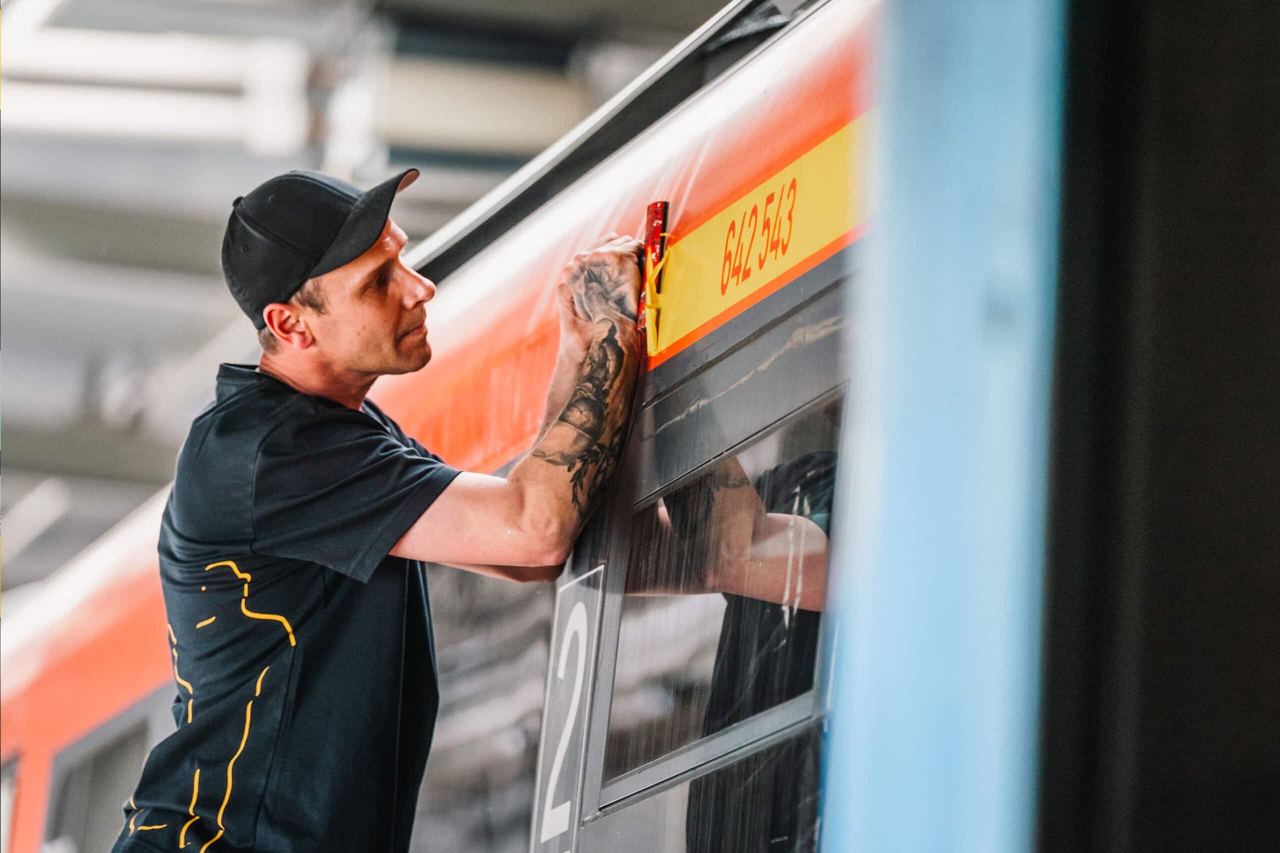 Ein Mitarbeiter entfernt mit großer Präzision eine Fahrzeugnummer auf einem Zug. Seine konzentrierte Haltung unterstreicht sein handwerkliches Können.