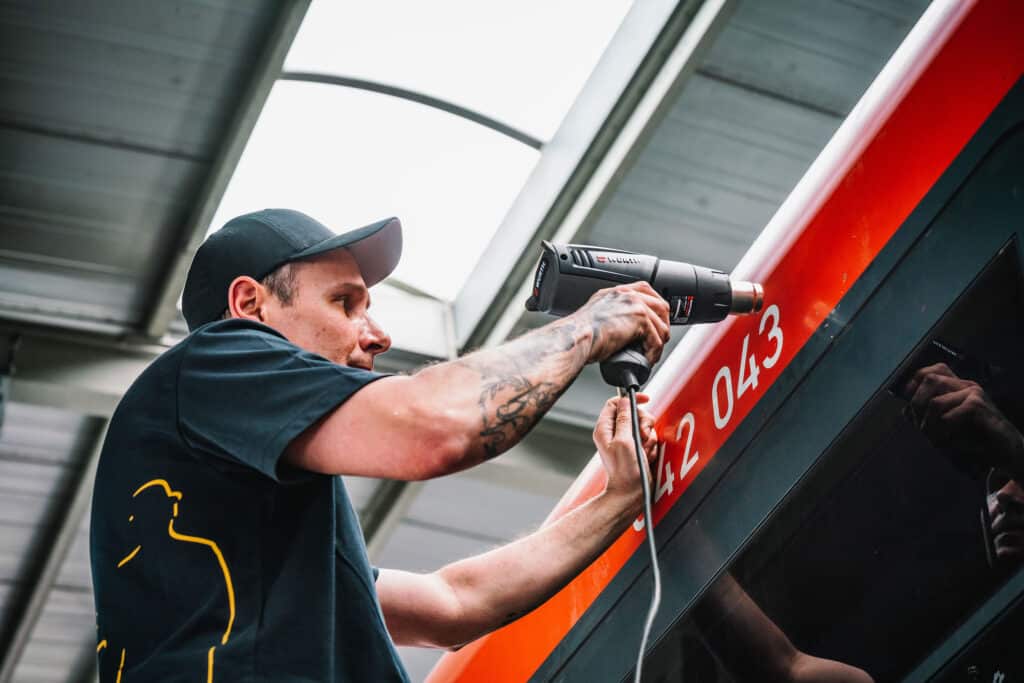 Ein Mitarbeiter erwärmt mit einer Heißluftpistole eine alte Beschriftung auf einem Fahrzeug, um sie leichter entfernen zu können. Mit konzentriertem Blick bereitet er die Oberfläche für die nächste Gestaltung vor.