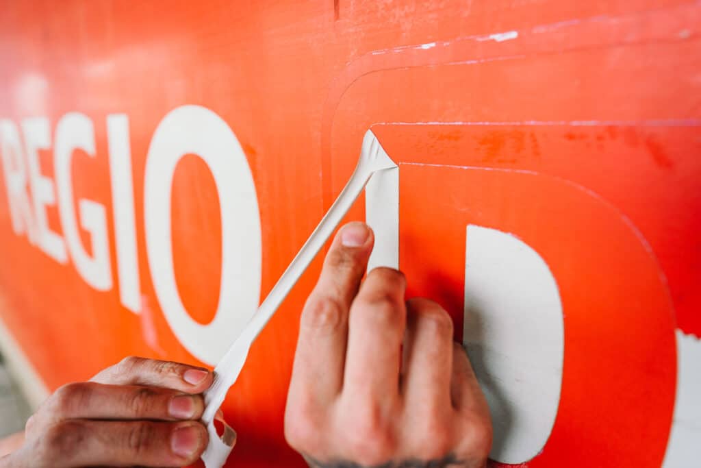 Ein Mitarbeiter entfernt präzise eine alte Beschriftung von einem Fahrzeug. Die weiße Folie wird von der orangefarbenen Oberfläche abgezogen, um Platz für eine neue Gestaltung zu schaffen.