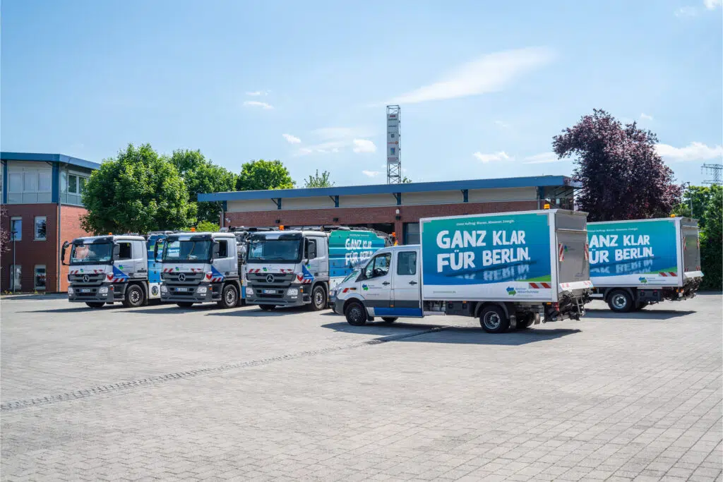 Mehrere Fahrzeuge der Berliner Wasserbetriebe auf einem Betriebshof. Im Vordergrund ein Transporter mit der Aufschrift „GANZ KLAR FÜR BERLIN.“ Dahinter LKWs mit Wasserbetriebe-Branding. Die Szene wird von blauem Himmel und modernen Gebäuden umrahmt.