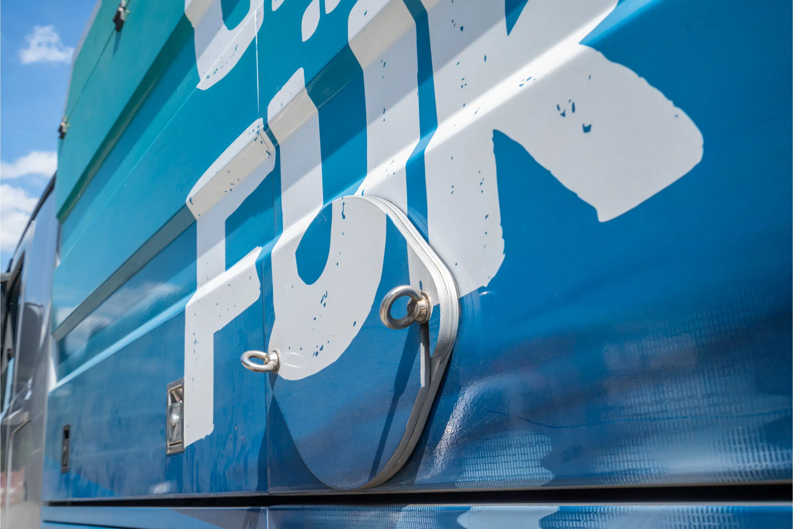 Nahaufnahme einer blauen Fahrzeugverkleidung mit weißer Schrift in verwittertem Design. Im Fokus steht eine runde, mit Metallösen gesicherte Wartungsklappe. Die glänzende Oberfläche reflektiert den Himmel und Umgebung, was dem Bild eine dynamische Optik verleiht.
