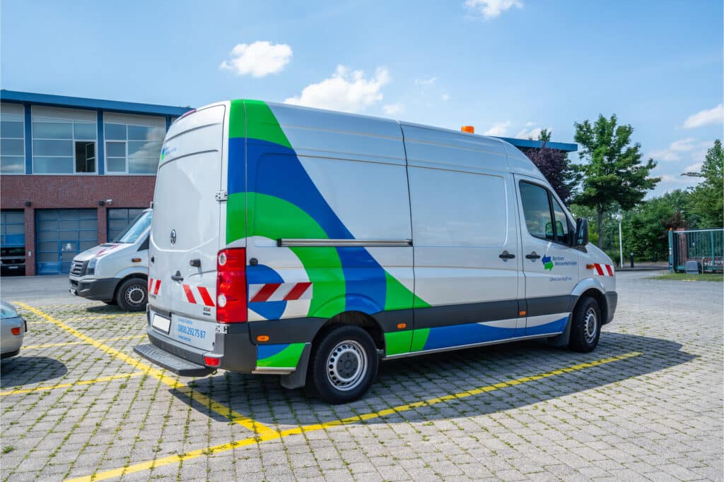 Ein silberner Transporter der Berliner Wasserbetriebe mit einer modernen, blau-grünen Folierung steht auf einem Parkplatz. Das Fahrzeug trägt reflektierende Warnmarkierungen und ein Dienstlogo. Im Hintergrund sind weitere Fahrzeuge und ein Firmengebäude zu sehen.
