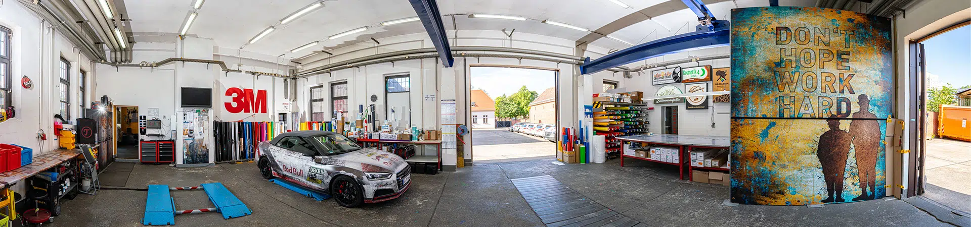 Das Bild zeigt eine Werkstatt mit einer offenen Garage. In der Mitte steht ein folierter Sportwagen mit Red-Bull-Design. An den Wänden befinden sich Regale mit bunten Folienrollen und Schildern. Rechts ist eine große Tür mit dem Schriftzug „DON’T HOPE WORK HARD“ und zwei Silhouetten.