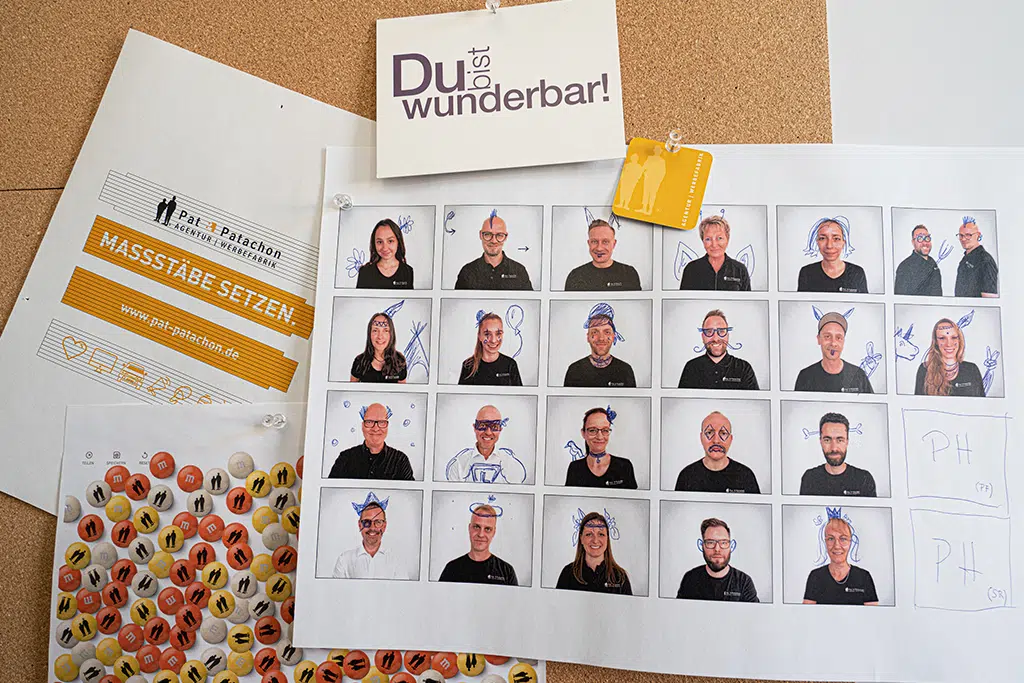 Eine Pinnwand mit einer Teamauswahl von Pat & Patachon. Einige Teamfotos sind kreativ mit blauen Zeichnungen versehen. Daneben ein Plakat mit "Maßstäbe setzen", eine Karte mit "Du bist wunderbar!" und individuell bedruckte M&M’s.