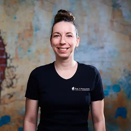 Eine Frau mit hochgesteckten Haaren trägt ein schwarzes T-Shirt mit Firmenlogo. Sie steht vor einer strukturierten Wand mit blauen und gelben Akzenten und lächelt in die Kamera.