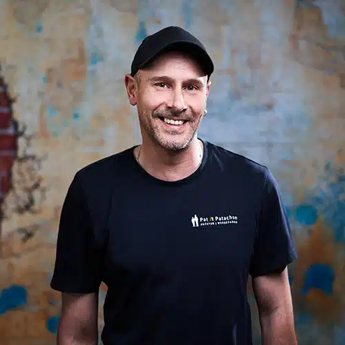 Ein Mann mit kurzem Bart und schwarzer Kappe trägt ein schwarzes T-Shirt mit dem Logo von Pat & Patachon. Er steht vor einer strukturierten Wand mit blauen und gelben Akzenten und lächelt freundlich in die Kamera.
