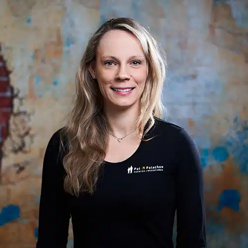 Eine Frau mit langen, welligen blonden Haaren trägt ein schwarzes Langarmshirt mit Firmenlogo. Sie steht vor einer strukturierten Wand mit blauen und gelben Akzenten und lächelt in die Kamera.