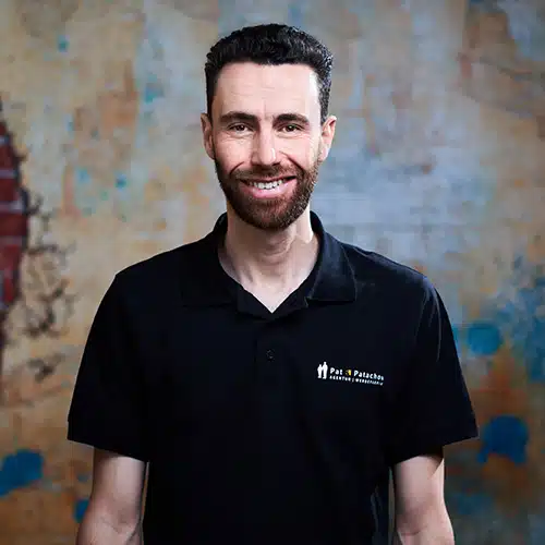 Ein Mann mit kurzen dunklen Haaren und Bart trägt ein schwarzes Poloshirt mit Firmenlogo. Er steht vor einer strukturierten Wand mit blauen und gelben Akzenten und lächelt in die Kamera.