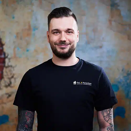 Ein Mann mit kurz rasierten Seiten und dunklem Bart trägt ein schwarzes T-Shirt mit Firmenlogo. Er hat tätowierte Arme und steht lächelnd vor einer Wand mit blauen und gelben Akzenten