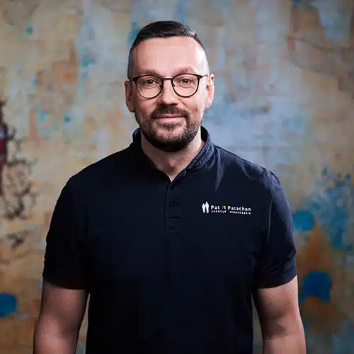Ein Mann mit Kurzhaarschnitt, Bart und Brille steht vor einer strukturierten, gemusterten Wand. Er trägt ein schwarzes Poloshirt mit dem Pat & Patachon-Logo. Sein Blick ist freundlich und direkt zur Kamera gerichtet.