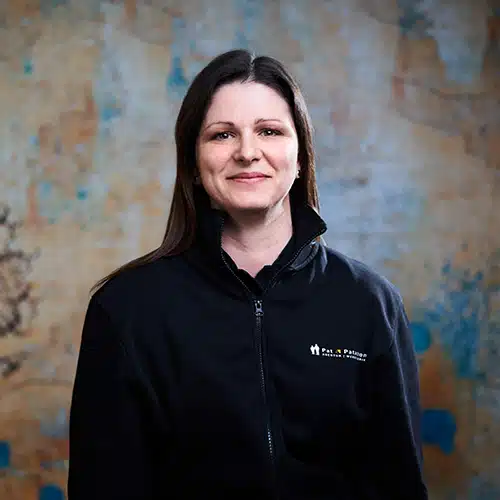 Eine Frau mit langen braunen Haaren trägt eine schwarze Jacke mit Firmenlogo. Sie steht vor einer strukturierten Wand mit blauen und gelben Akzenten und lächelt freundlich in die Kamera.