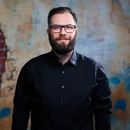 Ein Mann mit dunklem Vollbart und Brille steht vor einer strukturierten Wand mit blau-gelben Akzenten. Er trägt ein schwarzes Hemd und blickt freundlich in die Kamera.