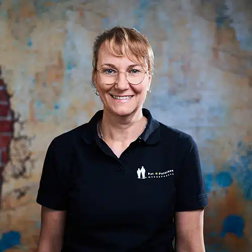 Eine Frau mit zusammengebundenen blonden Haaren und Brille trägt ein schwarzes Poloshirt mit Firmenlogo. Sie steht vor einer strukturierten Wand mit blauen und gelben Akzenten und lächelt freundlich in die Kamera.