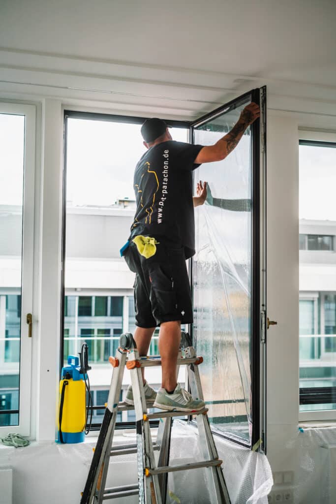 Ein Werbetechniker steht auf einer Leiter und bringt eine Schutzfolie auf eine große Fensterfläche auf. Er trägt schwarze Arbeitskleidung mit dem Logo von Pat & Patachon auf dem Rücken. Mit beiden Händen streicht er die Folie glatt, während sich Seifenwasser zwischen Glas und Folie verteilt. Eine Sprühflasche zur Befeuchtung steht auf der Fensterbank. Im Hintergrund sind moderne Bürogebäude zu sehen.