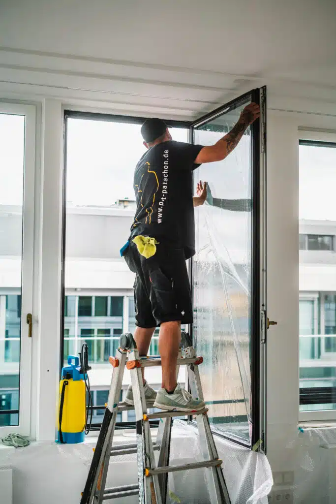 Ein Werbetechniker steht auf einer Leiter und bringt eine Schutzfolie auf eine große Fensterfläche auf. Er trägt schwarze Arbeitskleidung mit dem Logo von Pat & Patachon auf dem Rücken. Mit beiden Händen streicht er die Folie glatt, während sich Seifenwasser zwischen Glas und Folie verteilt. Eine Sprühflasche zur Befeuchtung steht auf der Fensterbank. Im Hintergrund sind moderne Bürogebäude zu sehen.