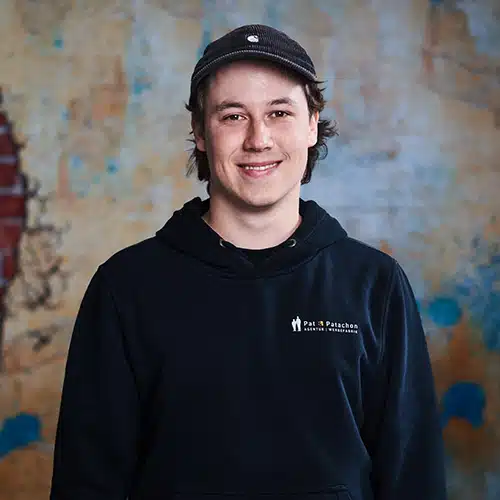 Porträt eines lächelnden Mannes mit kinnlangen, braunen leicht lockigen Haaren. Er trägt einen schwarzen Hoodie mit Pat & Patachon Aufdruck und ein Cap. Er steht vor einer künstlerisch gestalteten Wand mit Backstein-Elementen in Form einer Silhouette. Die warme, strukturierte Hintergrundgestaltung verleiht dem Bild eine moderne und kreative Atmosphäre.