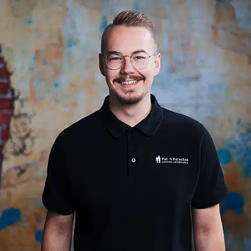 Porträt eines lächelnden Mannes mit kurzen, blond gestrichenen Haaren und Bart. Er trägt eine Brille und ein schwarzes Poloshirt mit Pat & Patachon Aufdruck. Er steht vor einer künstlerisch gestalteten Wand mit Backstein-Elementen in Form einer Silhouette. Die warme, strukturierte Hintergrundgestaltung verleiht dem Bild eine moderne und kreative Atmosphäre.
