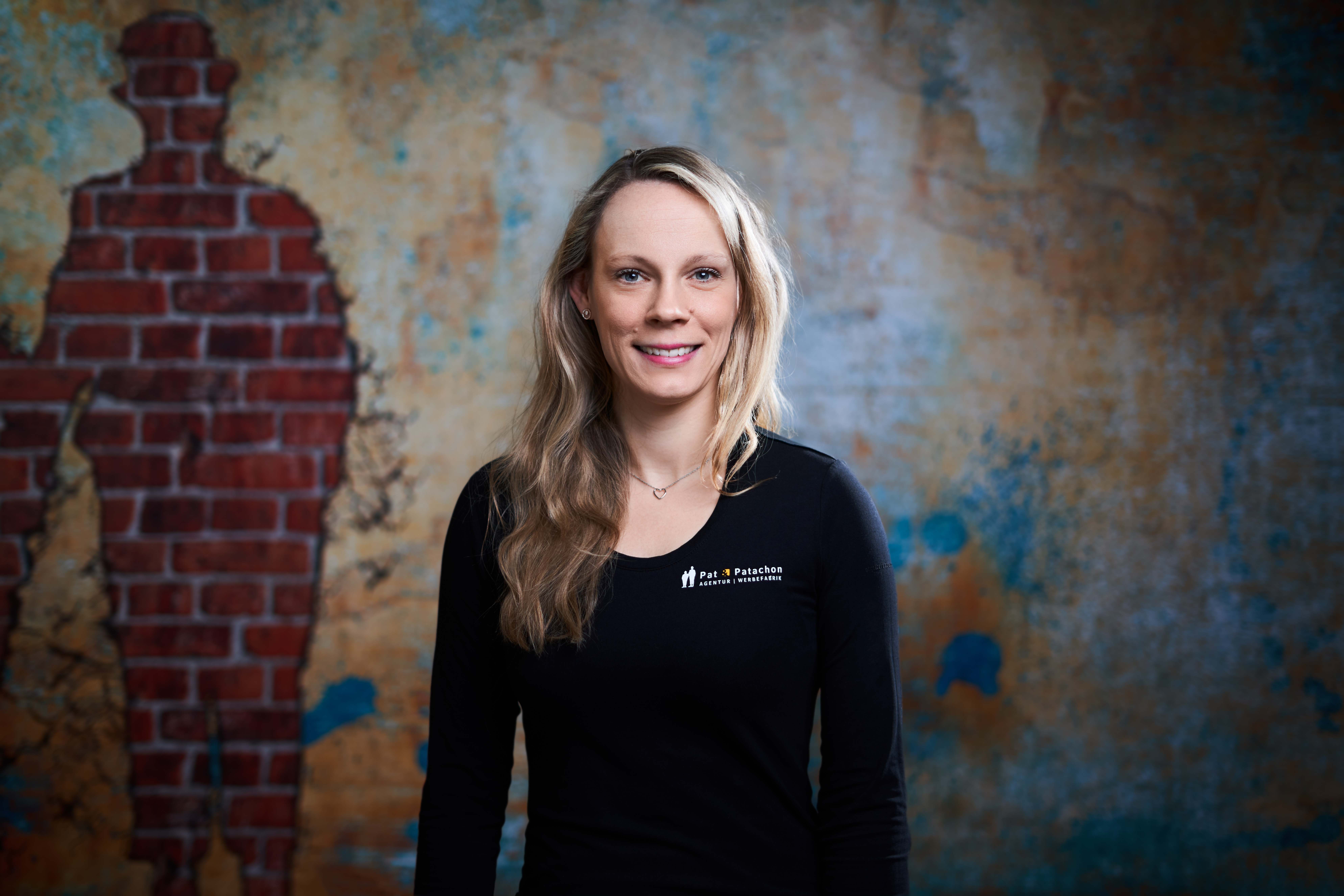 Porträt einer lächelnden Frau mit langen, blonden Haaren, welche auf ihrer Schulter liegen. Sie trägt ein schwarzes Langarmshirt mit dem Aufdruck "Pat & Patachon" und steht vor einer künstlerisch gestalteten Wand mit Backstein-Elementen in Form einer Silhouette. Die warme, strukturierte Hintergrundgestaltung verleiht dem Bild eine moderne und kreative Atmosphäre.