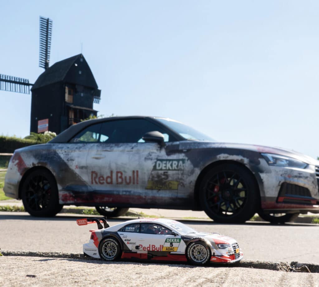 Ein im Motorsport-Look foliertes Auto mit Red Bull Branding steht auf einer Straße vor einer traditionellen Windmühle. Im Vordergrund befindet sich ein ferngesteuertes Modellauto im gleichen Design, was eine beeindruckende Perspektive und einen kreativen Größenvergleich schafft.