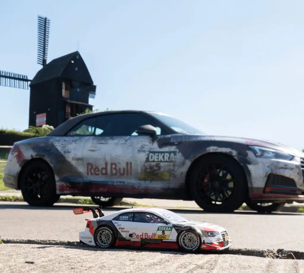 Ein im Motorsport-Look foliertes Auto mit Red Bull Branding steht auf einer Straße vor einer traditionellen Windmühle. Im Vordergrund befindet sich ein ferngesteuertes Modellauto im gleichen Design, was eine beeindruckende Perspektive und einen kreativen Größenvergleich schafft.