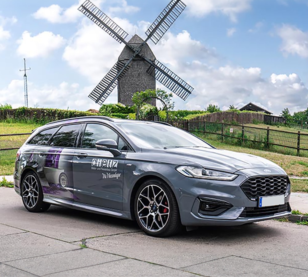 Ein stilvoll foliertes Auto mit Branding für 'dietz' steht auf einer gepflasterten Fläche vor einer traditionellen Windmühle. Die Fahrzeugbeschriftung integriert ein technisches Motiv mit lila Akzenten auf dem grauen Lack, während das Auto sportliche Felgen und rote Bremssättel trägt. Der Himmel ist leicht bewölkt, und die grüne Landschaft im Hintergrund rundet das Bild ab.