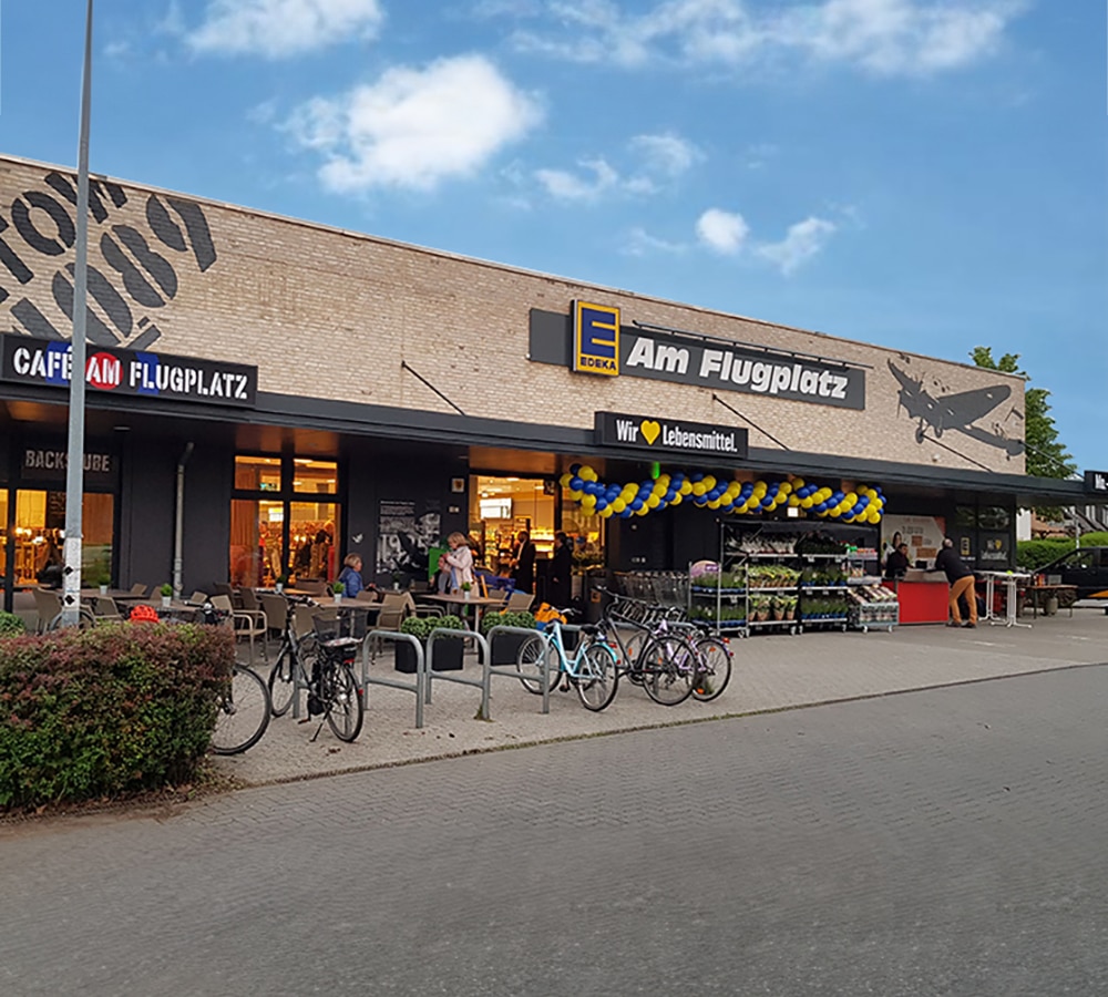 Der Eingangsbereich eines Edeka-Supermarkts mit der Aufschrift 'Am Flugplatz'. Das Gebäude hat eine helle Backsteinfassade mit einem aufgedruckten Flugzeugmotiv und Schriftzügen. Über dem Eingang hängt eine blau-gelbe Girlande aus Luftballons. Davor befinden sich Fahrradständer mit abgestellten Fahrrädern, Sitzplätze eines Cafés mit Gästen sowie eine Auslage mit Pflanzen. Der Himmel ist leicht bewölkt, und die Szenerie wirkt einladend und geschäftig.