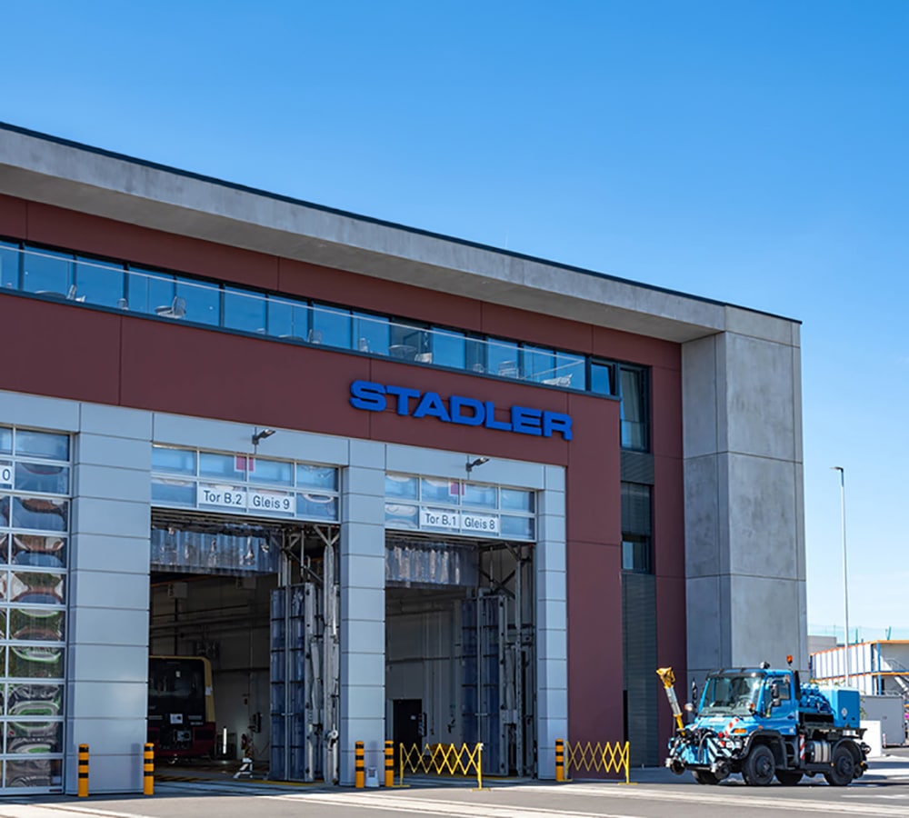 Moderne Industriehalle mit großem 'STADLER'-Firmenschild in blauen 3D-Buchstaben an der Fassade. Die Halle hat mehrere große, offene Tore mit der Beschriftung der Gleise, wodurch ein Zug im Inneren sichtbar ist. Im Vordergrund steht ein blaues Spezialfahrzeug. Der Himmel ist klar und blau, was die moderne Architektur und das klare Design des Gebäudes betont.