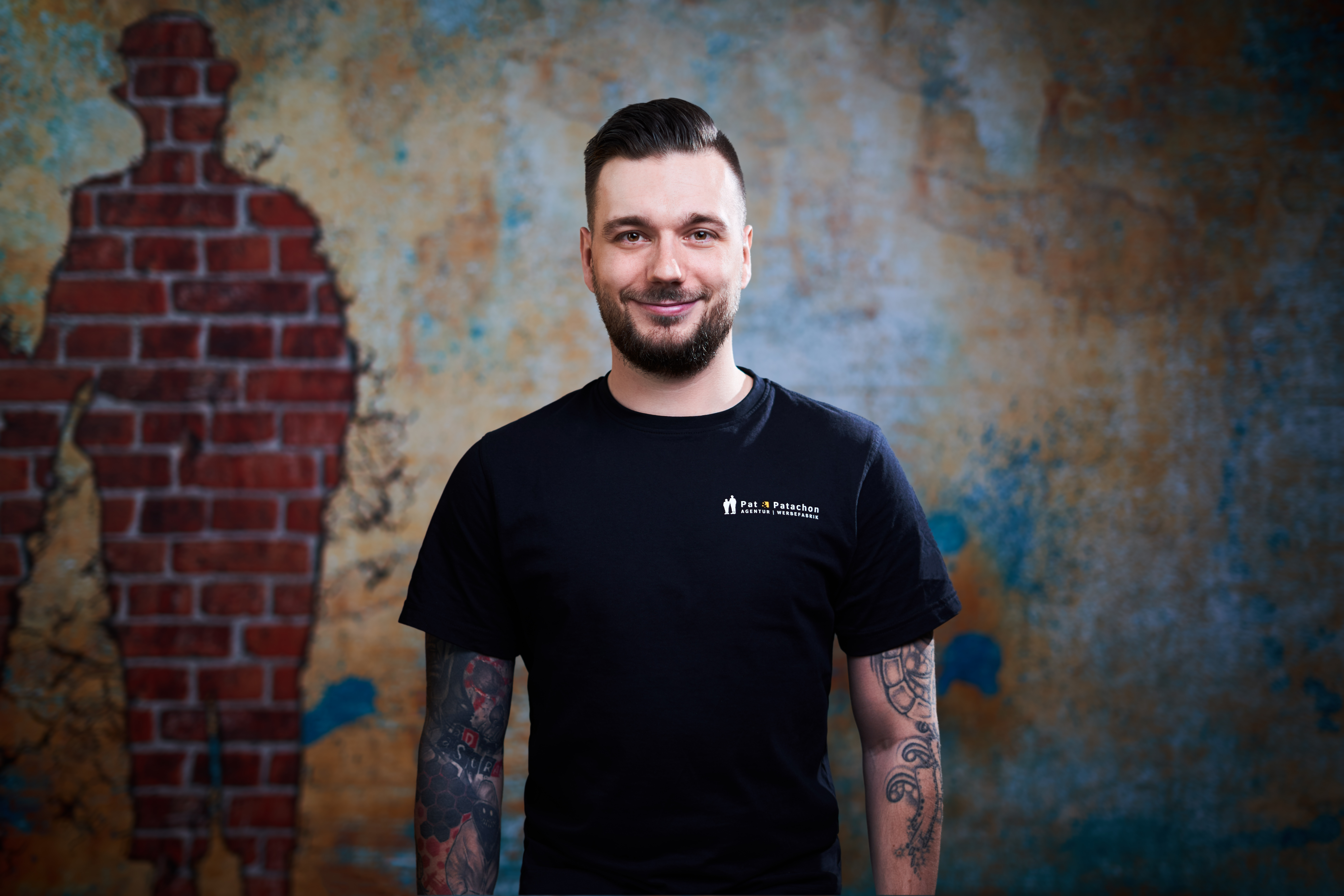 Ein freundlicher Mann mit dunklem Haar und Bart steht vor einer künstlerischen Wand mit Backstein-Silhouette. Er trägt ein schwarzes T-Shirt mit dem Logo von Pat & Patachon und hat tätowierte Arme. Sein Lächeln und die entspannte Haltung verleihen dem Bild eine positive und professionelle Ausstrahlung.